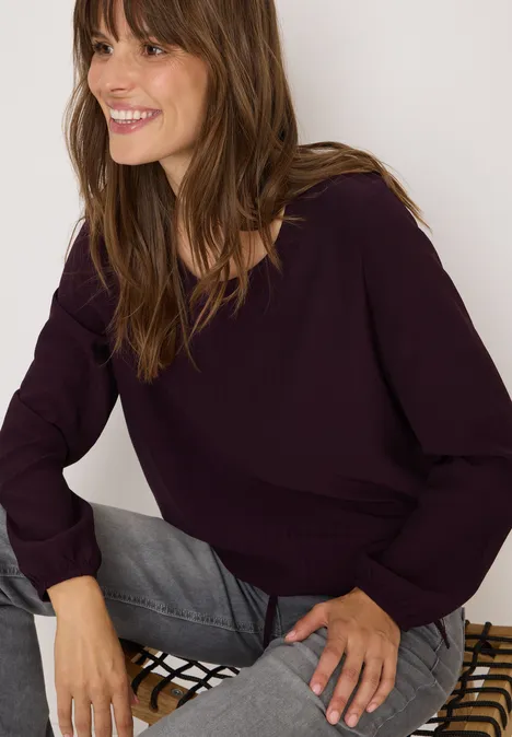 Frau mit lila Pullover und grauer Jeans sitzt und lächelt.